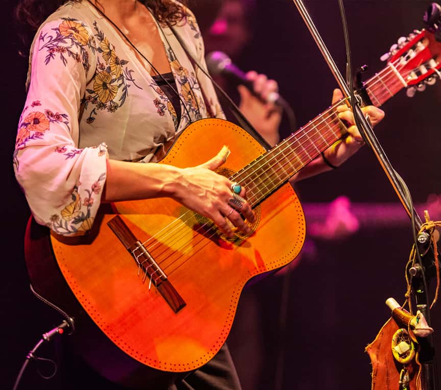 A musician plays an acoustic guitar on stage, wearing a floral blouse.