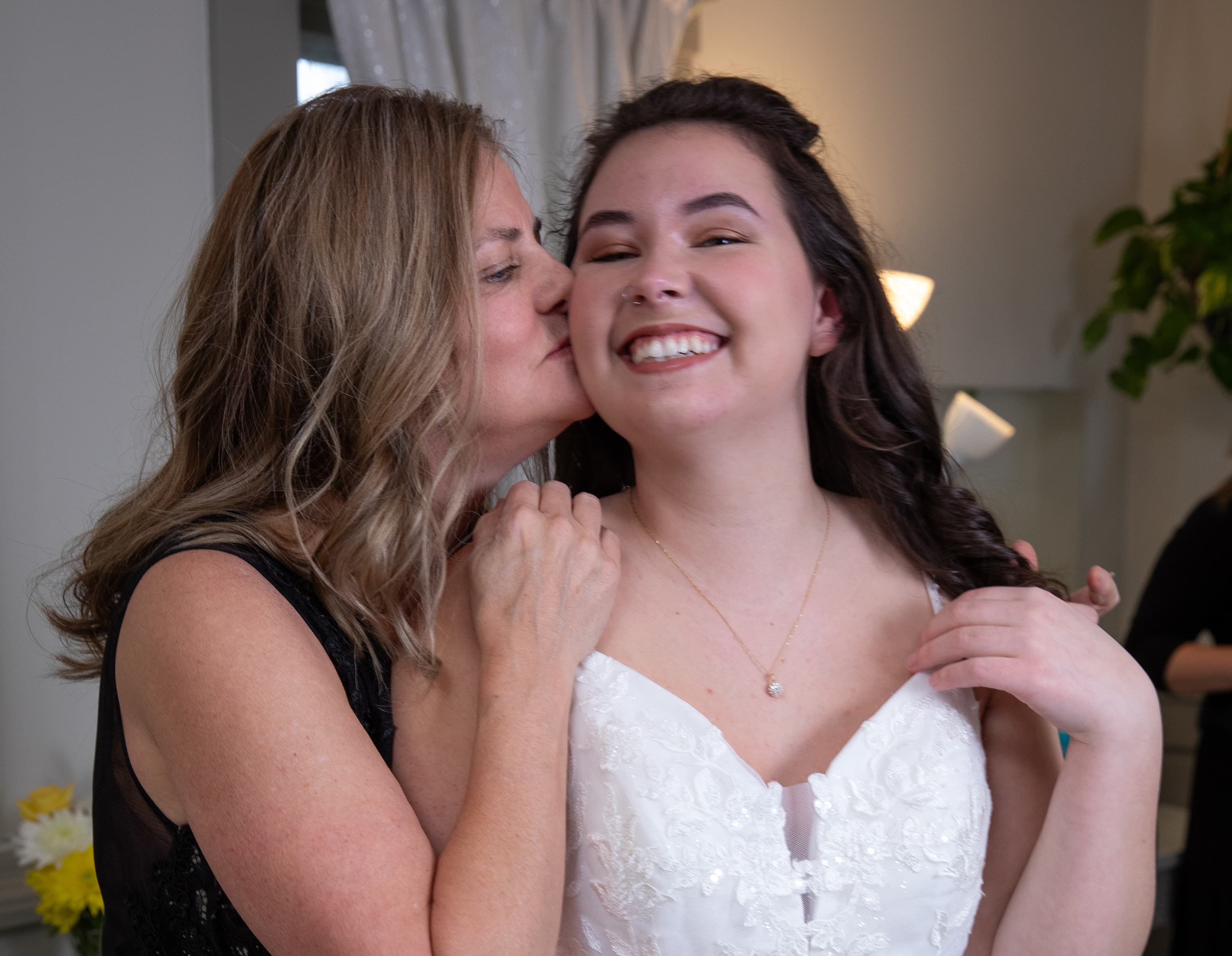A smiling bride gets a kiss from her mother in a bright room.