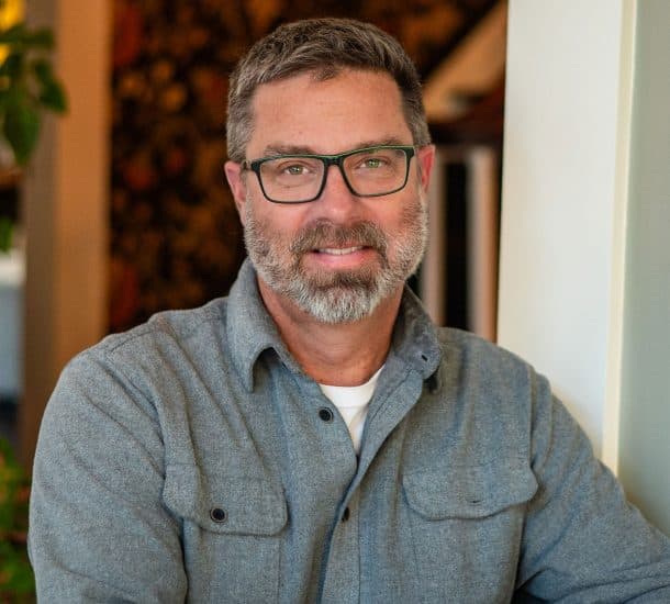 A man with glasses and a beard smiles while leaning against a wall in a cozy indoor setting.