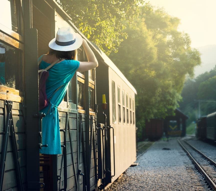 A person in a blue dress and white hat leans out of a train window, looking at the scenery.