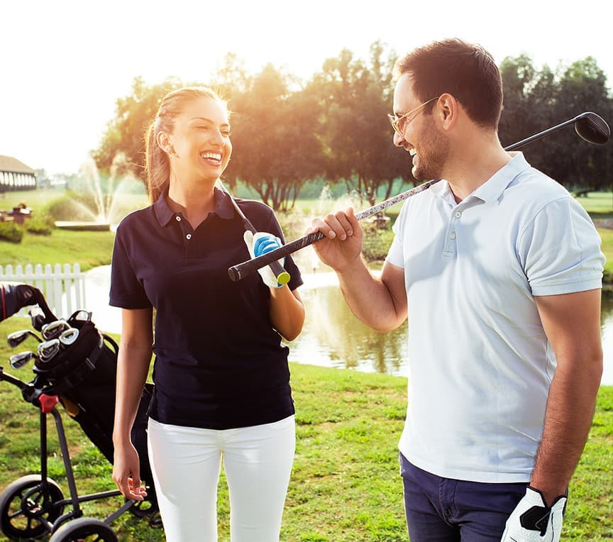 A man and woman are smiling and chatting while holding golf clubs on a sunny day near a pond.