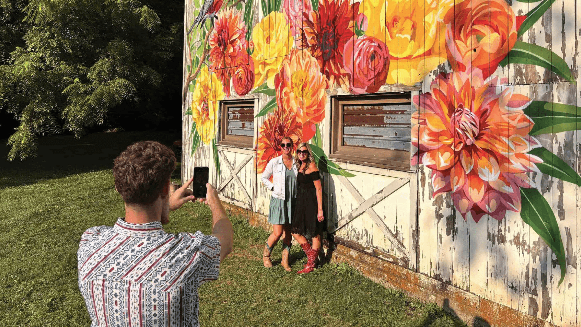 Two women pose for a photo in front of a colorful floral mural while a person takes their picture.