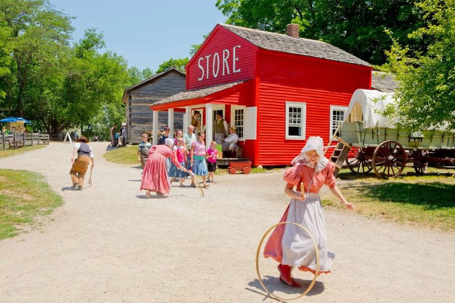 A red historic store building with visitors and children playing in a village setting. A red historic store building with visitors and children playing in a village setting.