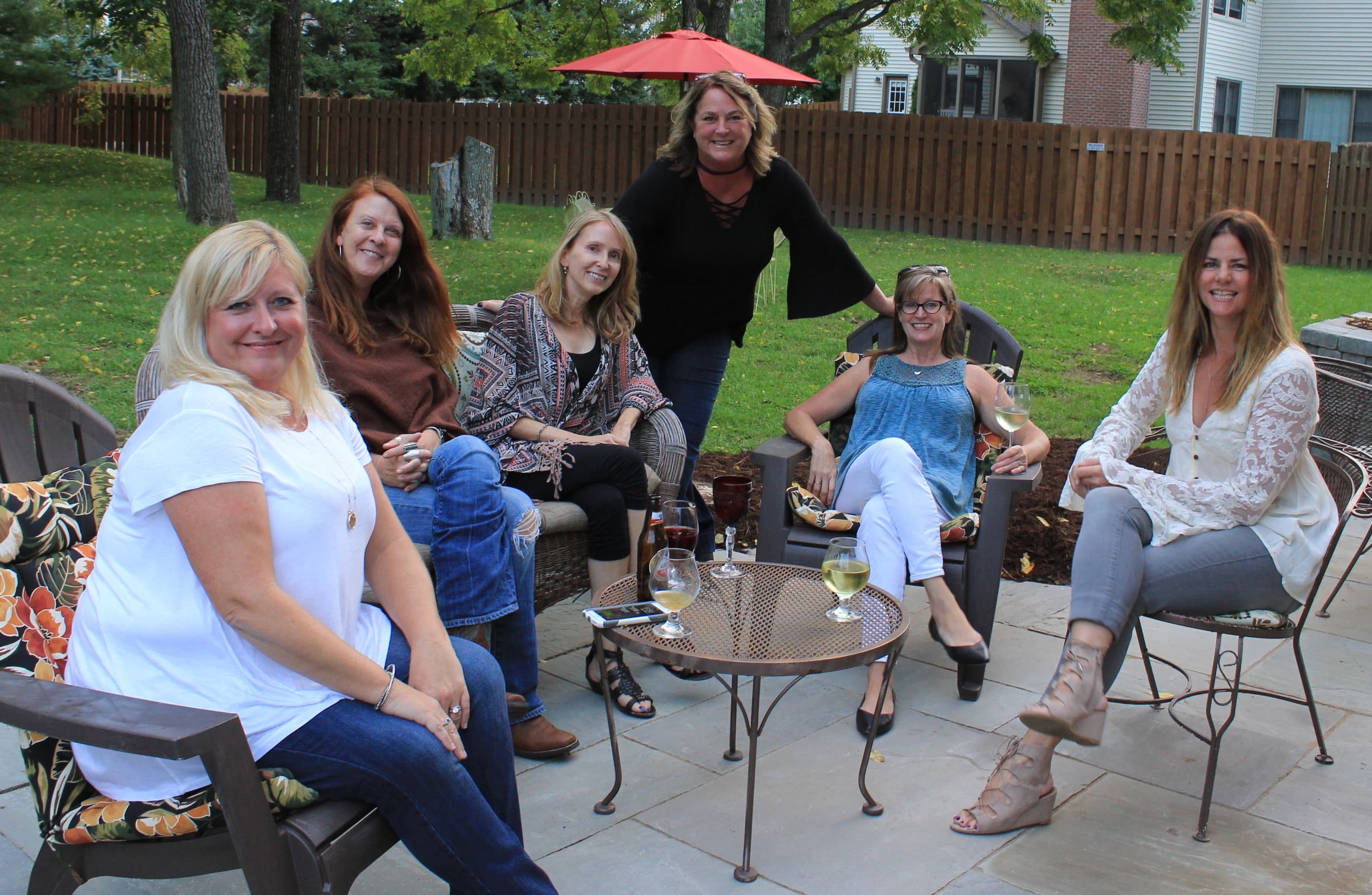 A group of six women sitting outdoors in a relaxed setting with drinks, smiling and enjoying each other's company.