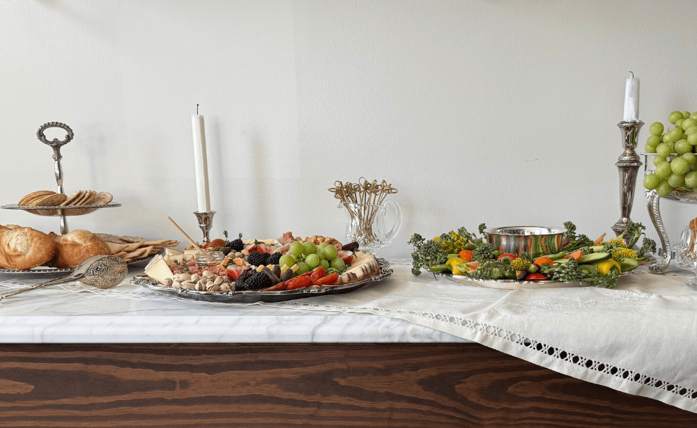 A beautifully arranged food spread featuring a cheese and fruit platter, pastries, and a vegetable display atop a white lace tablecloth.