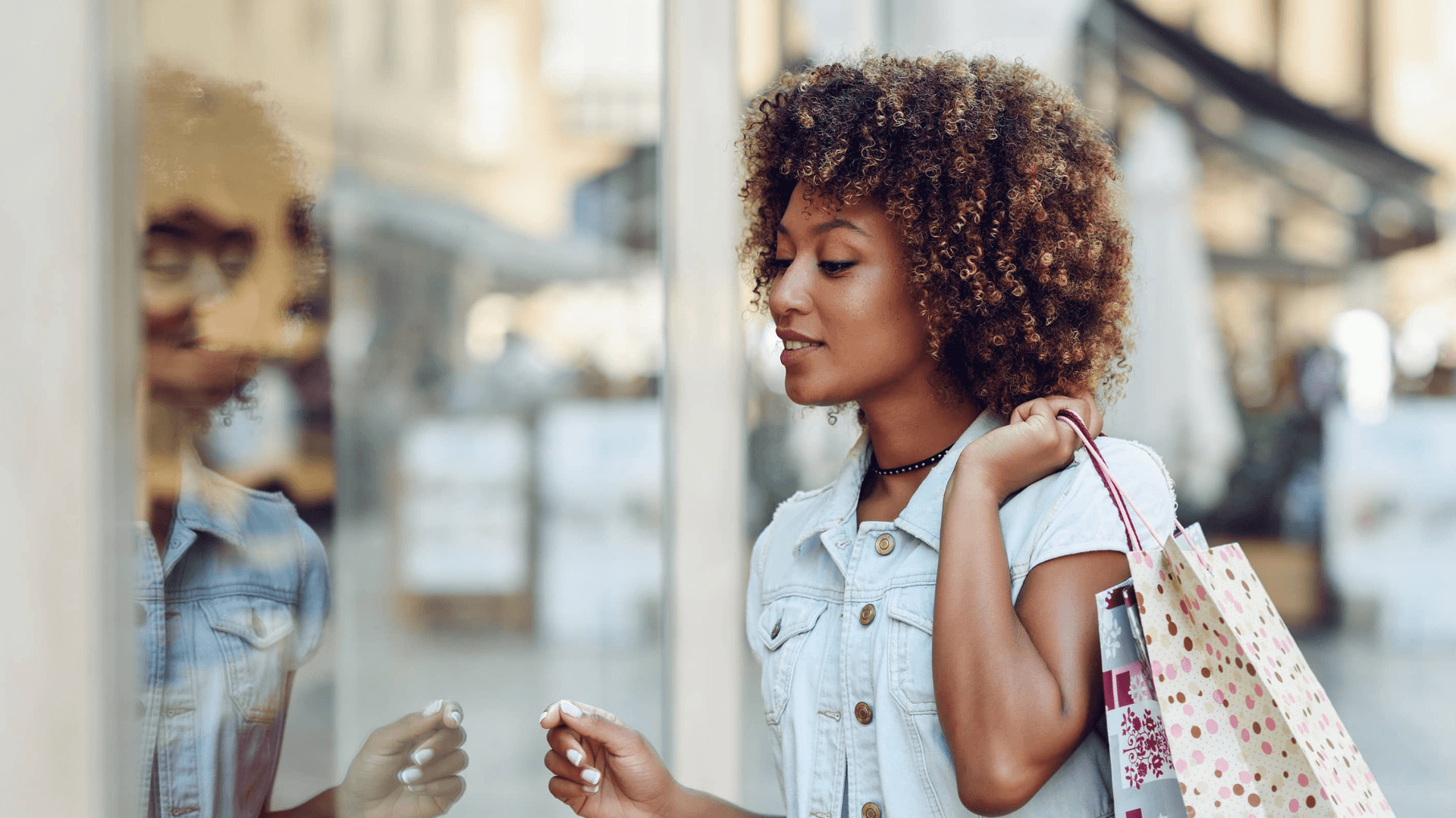 A woman with curly hair looks at herself in a shop window while holding shopping bags.