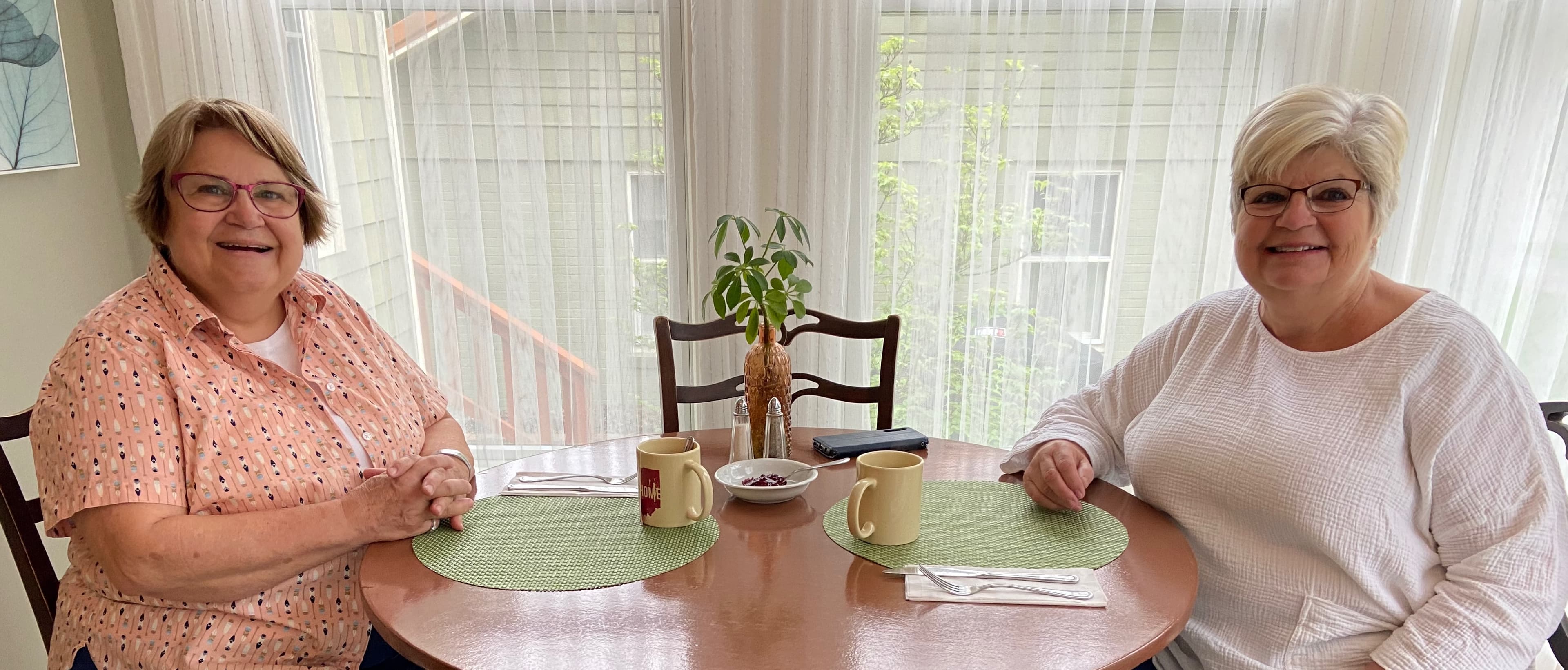 Two women sit at a table with green placemats, mugs, and a small plant in a sunlit room.