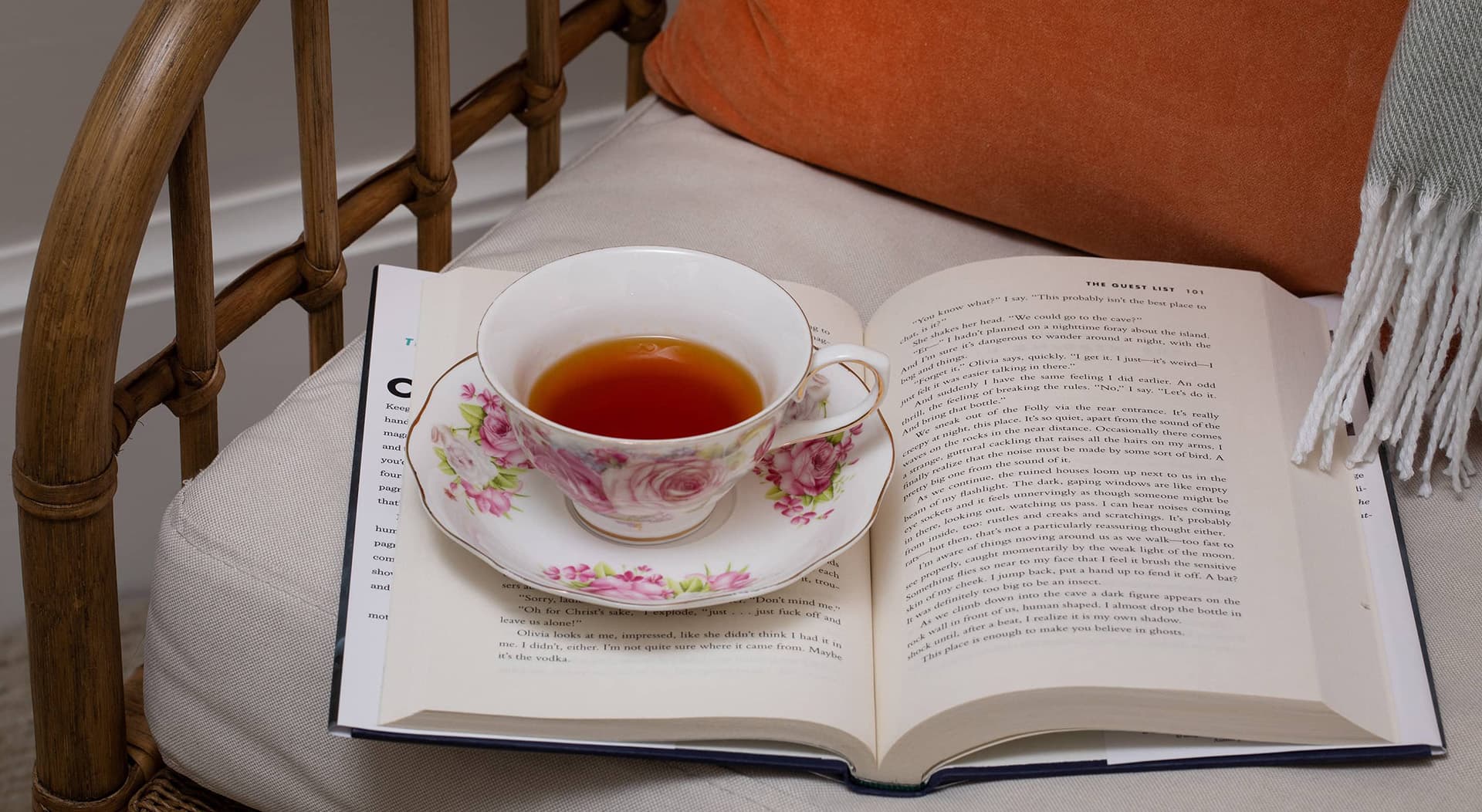 A delicate floral teacup rests on an open book, with a cozy chair and throw pillow in the background.