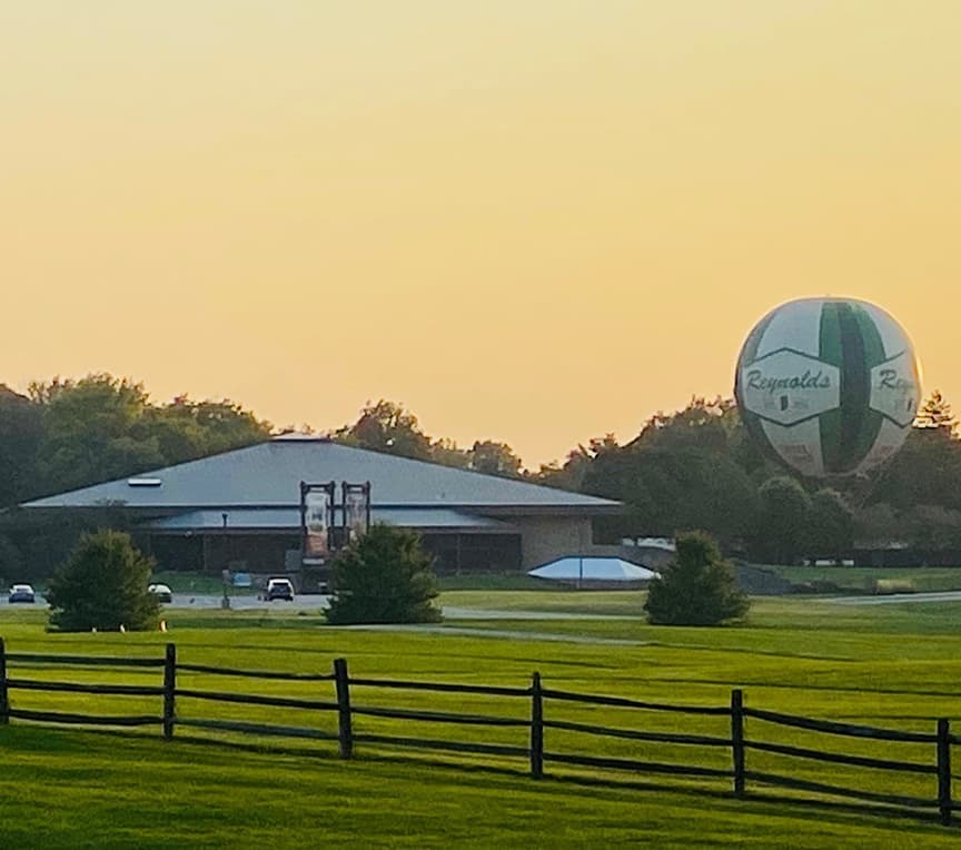 A large green and white hot air balloon floats above a building and grassy field at sunset.